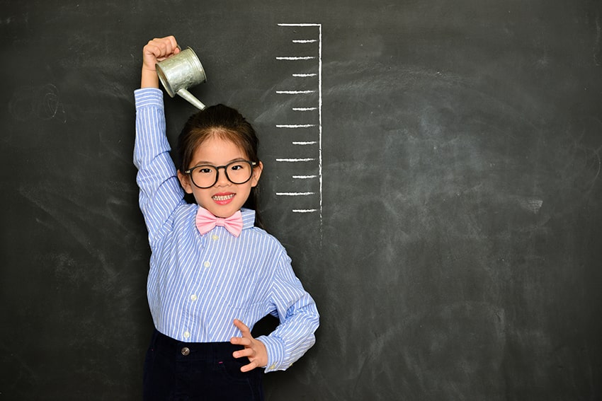 young asian girl with watering can stands in front of a blackboard with grown chart